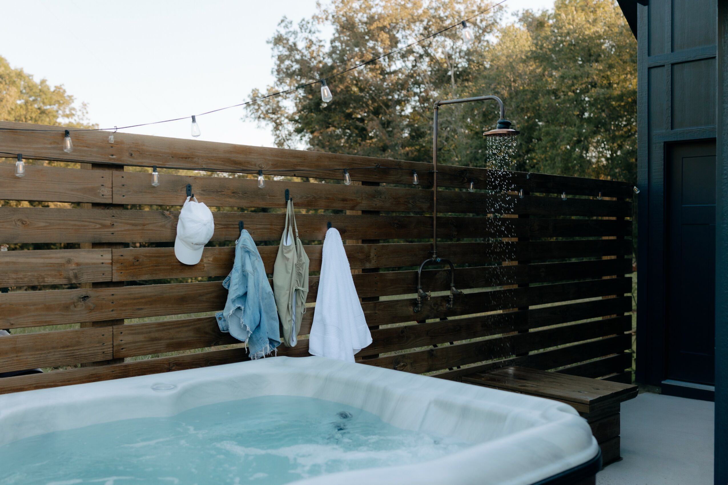 Outdoor hot tub with water, wooden fence, hanging towels, and showerhead, with trees in background.
