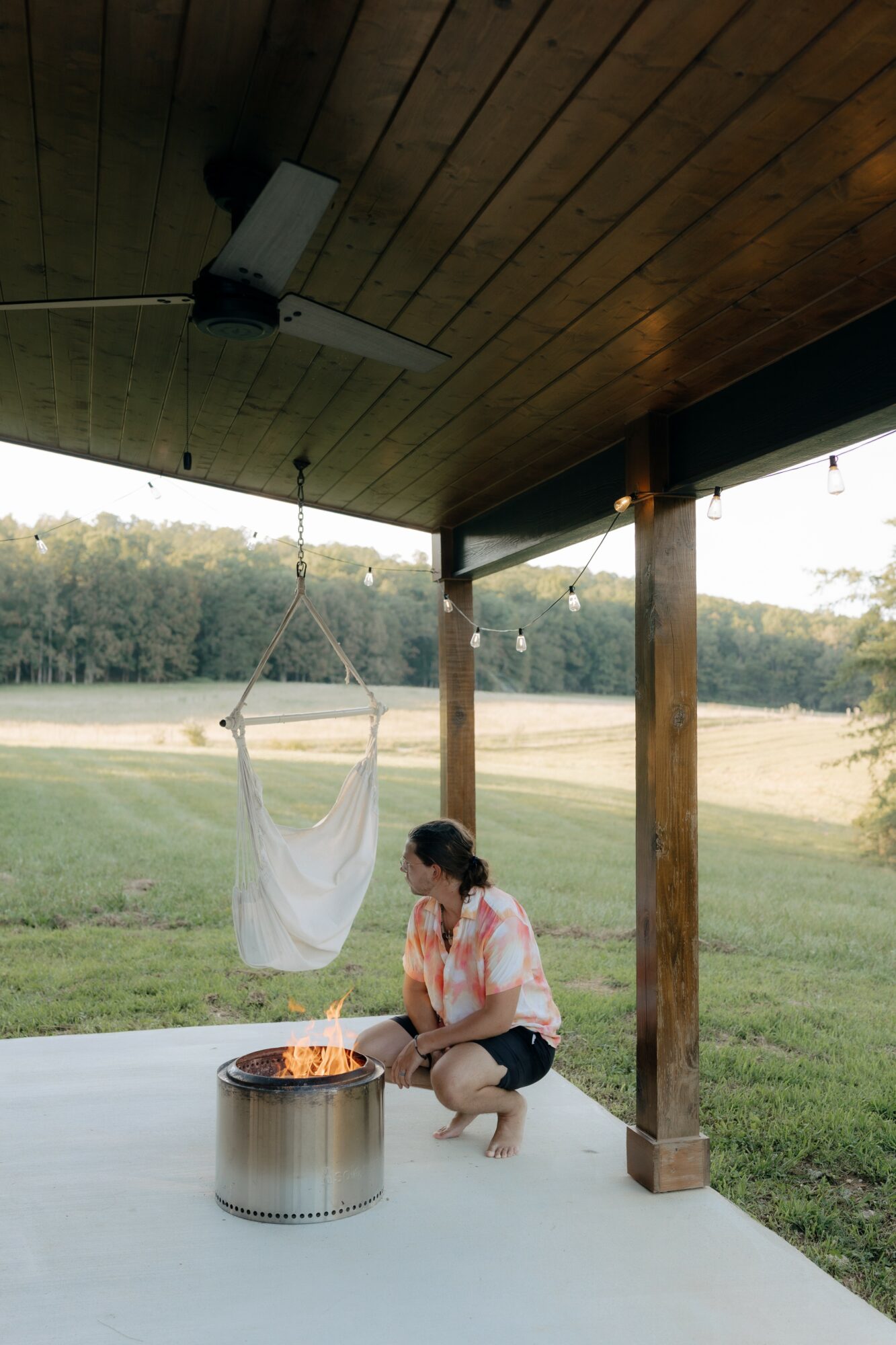 Person crouching near a fire pit on a porch with a wooden ceiling and support beams, overlooking a field and trees.