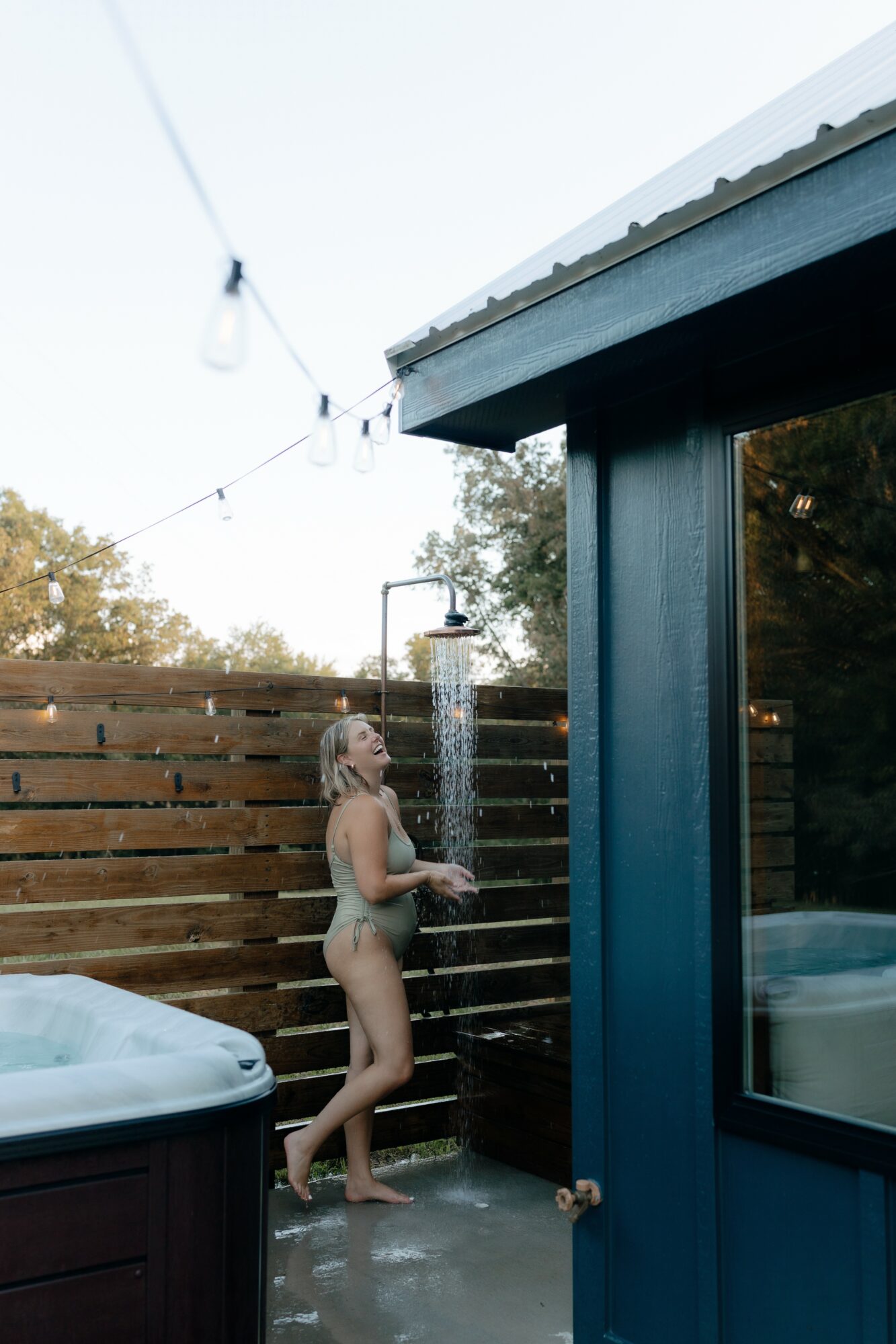 Woman standing outdoors near a shower with water flowing, wooden fence in background, outdoor setting.