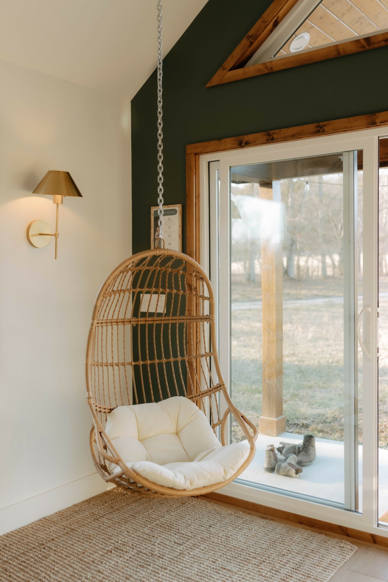 Wicker hanging chair with cushion near sliding glass door, outdoor view with trees and grass, indoor wall lamp, and ceiling window.