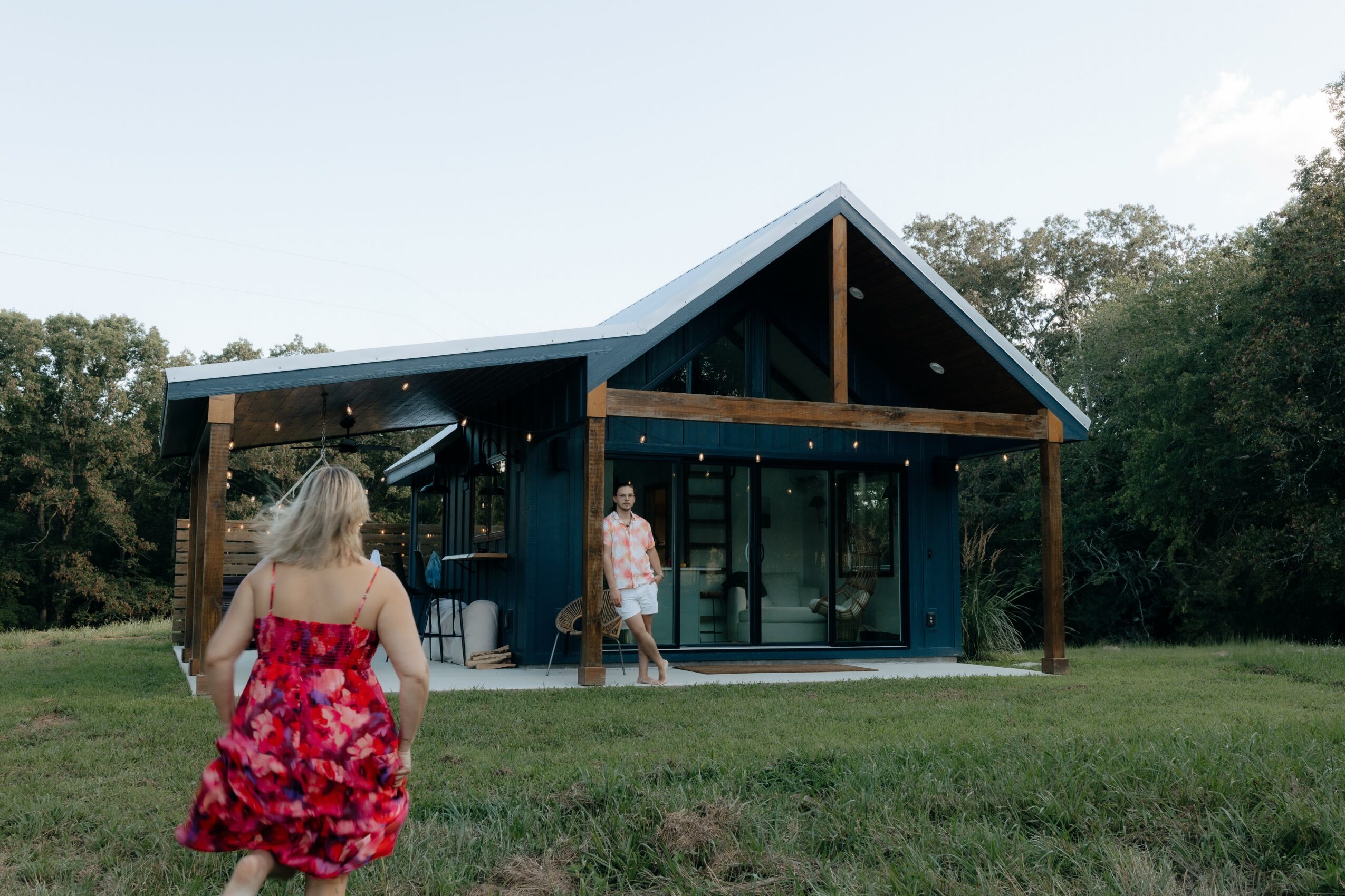 Child in a red floral dress walking towards a modern house with a triangular roof, glass walls, and wooden supports, outdoors.