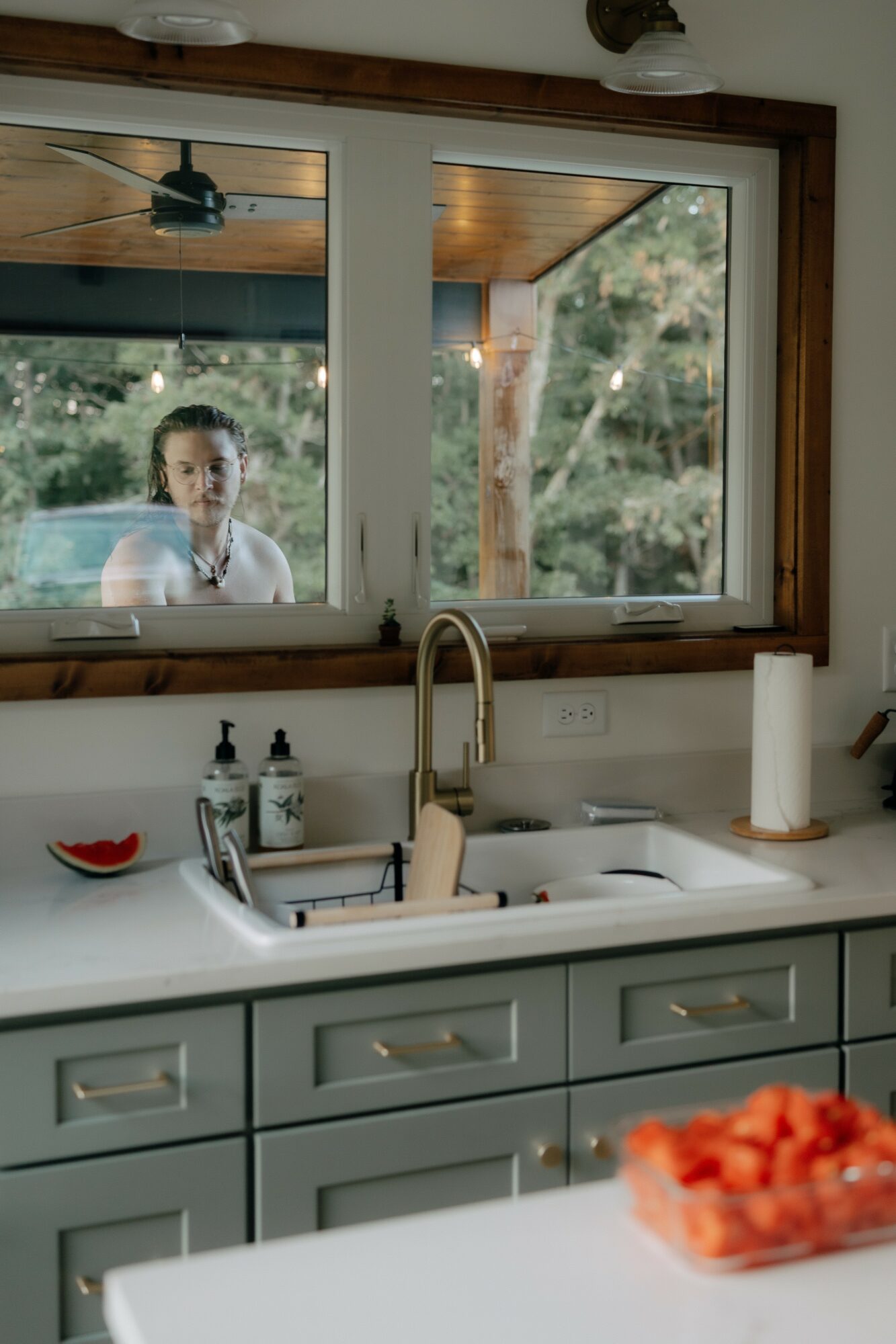 Kitchen sink with faucet, soap dispenser, and paper towel holder, viewed through a window showing a person outside.