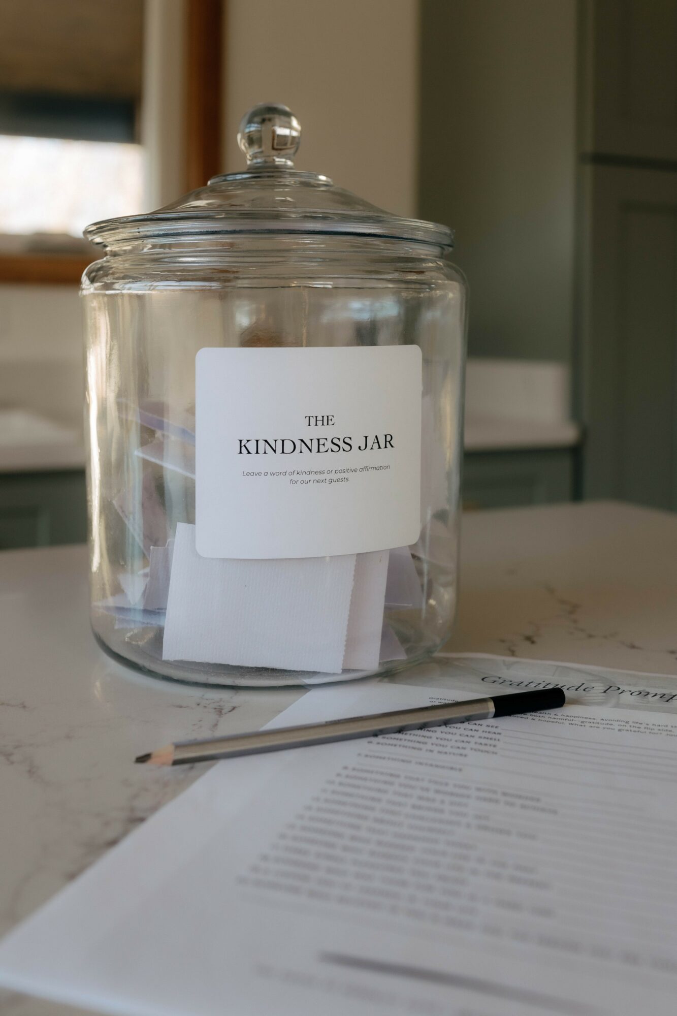 Glass jar with a lid, labeled 'The Kindness Jar,' on a table with papers and a pen nearby.