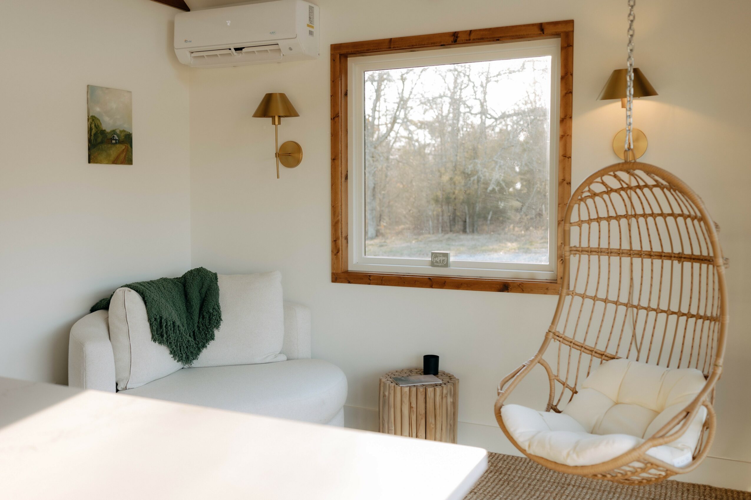 Living room with white sofa, hanging chair, window, and wall-mounted lights, decorated in neutral tones with natural light.