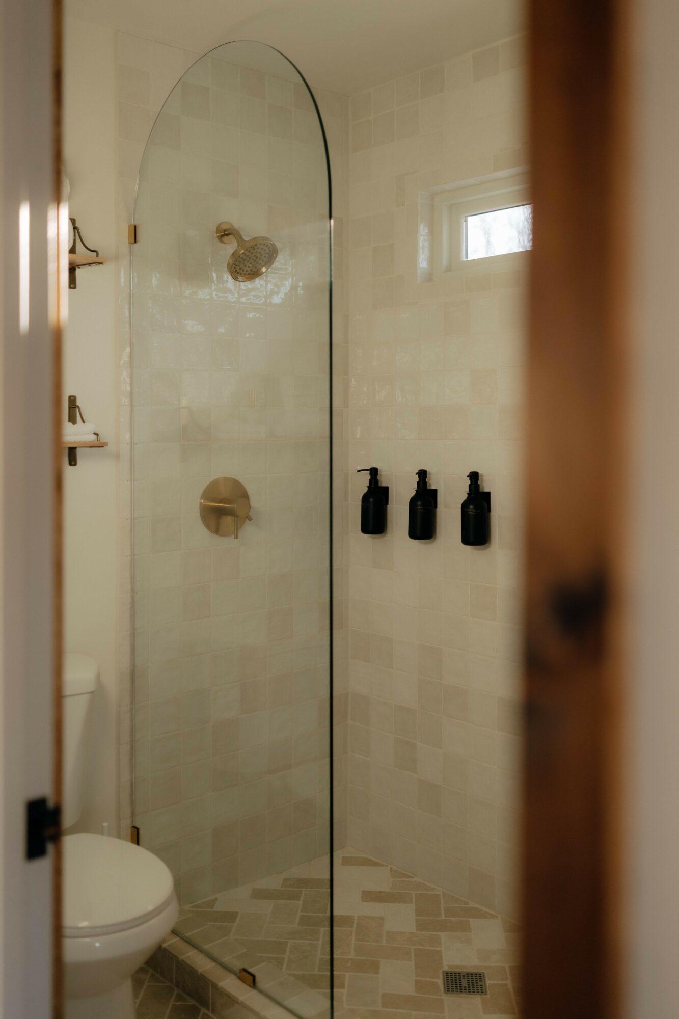 Bathroom shower with glass door, shelves, and three black bottles on wall, toilet visible in corner, window in background.