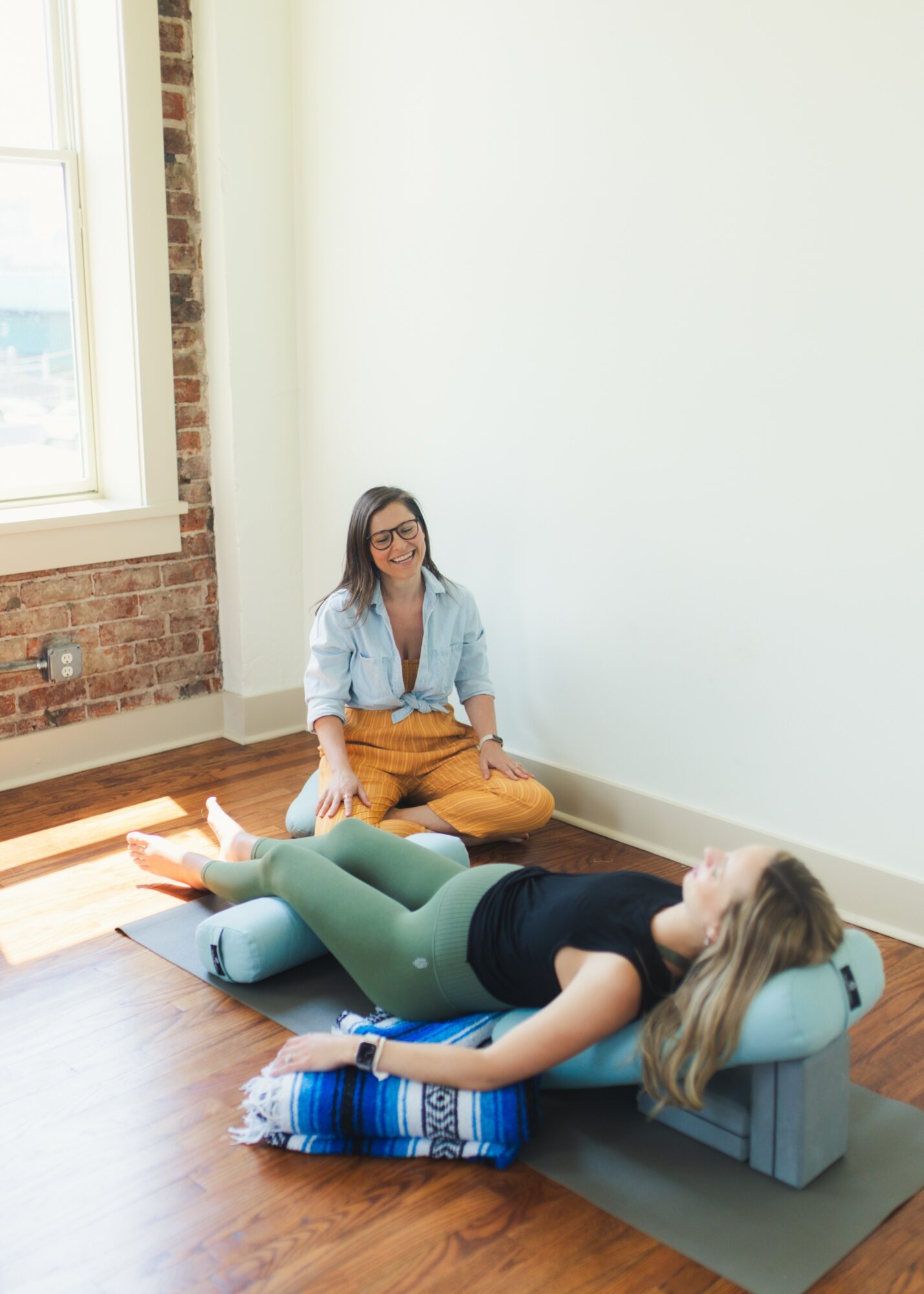 Two women in a bright room, one sitting on the floor and the other lying on a cushion, relaxing.