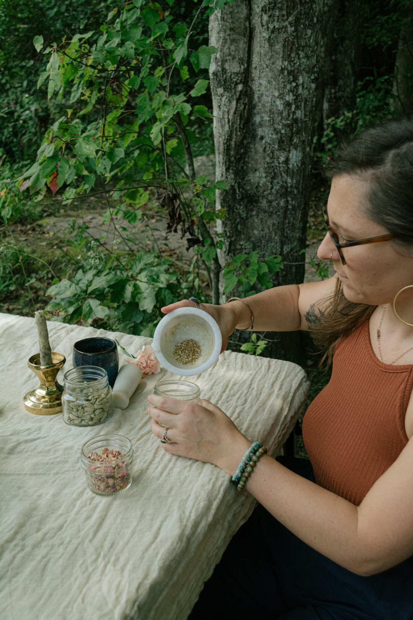 Woman with glasses sitting at a table outdoors, pouring a drink from a cup, surrounded by greenery.