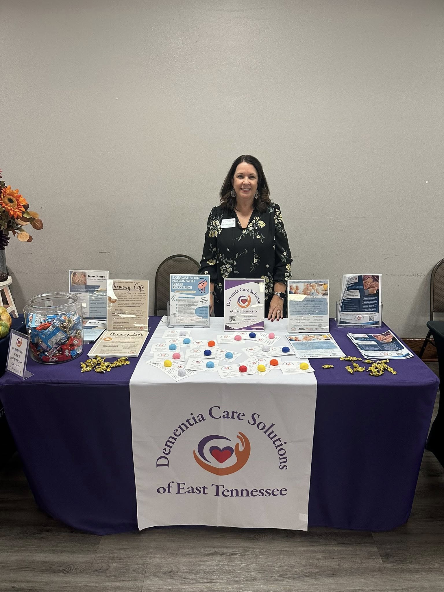 Woman standing behind a table with promotional materials, a banner reads 'Dementia Care Solutions of East Tennessee,' in a room with a gray wall.