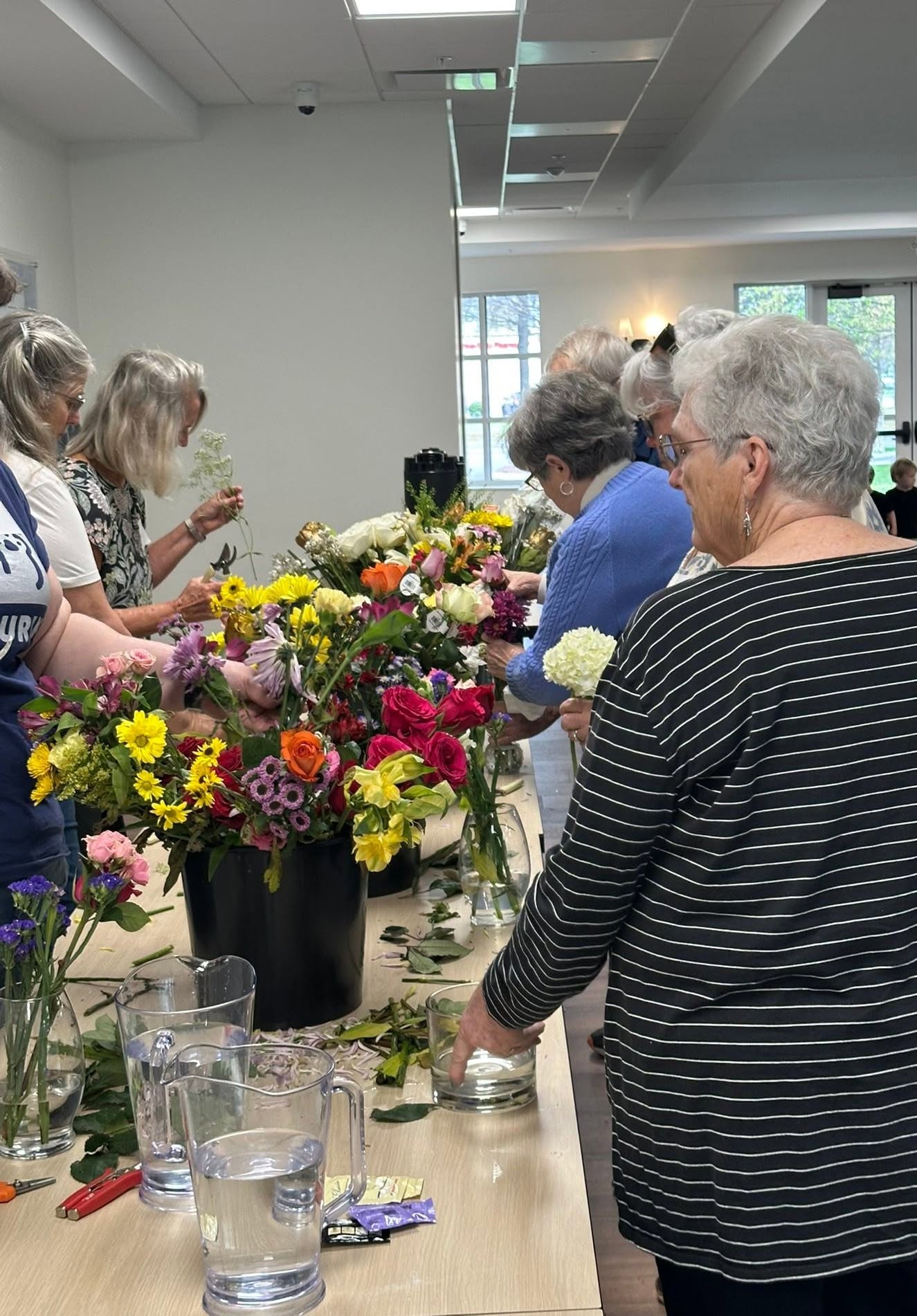 Group of women gathered around a table with colorful flowers, some holding flowers, in a bright indoor space.