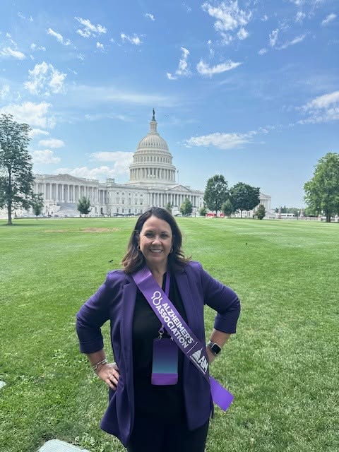 Woman standing on grass in front of the U.S. Capitol building, smiling, wearing a purple sash and jacket.