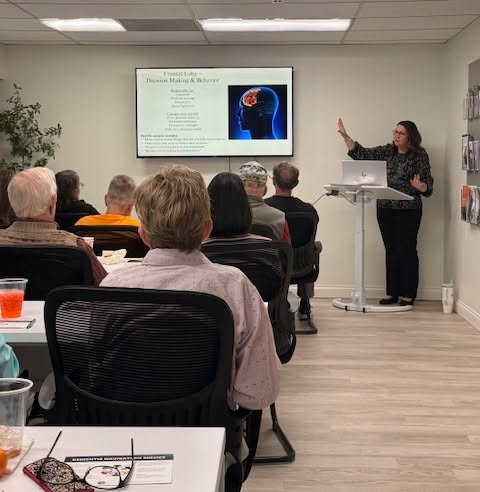 A woman presents a lecture to an audience in a room with a screen displaying a brain image and text, with attendees seated facing her.