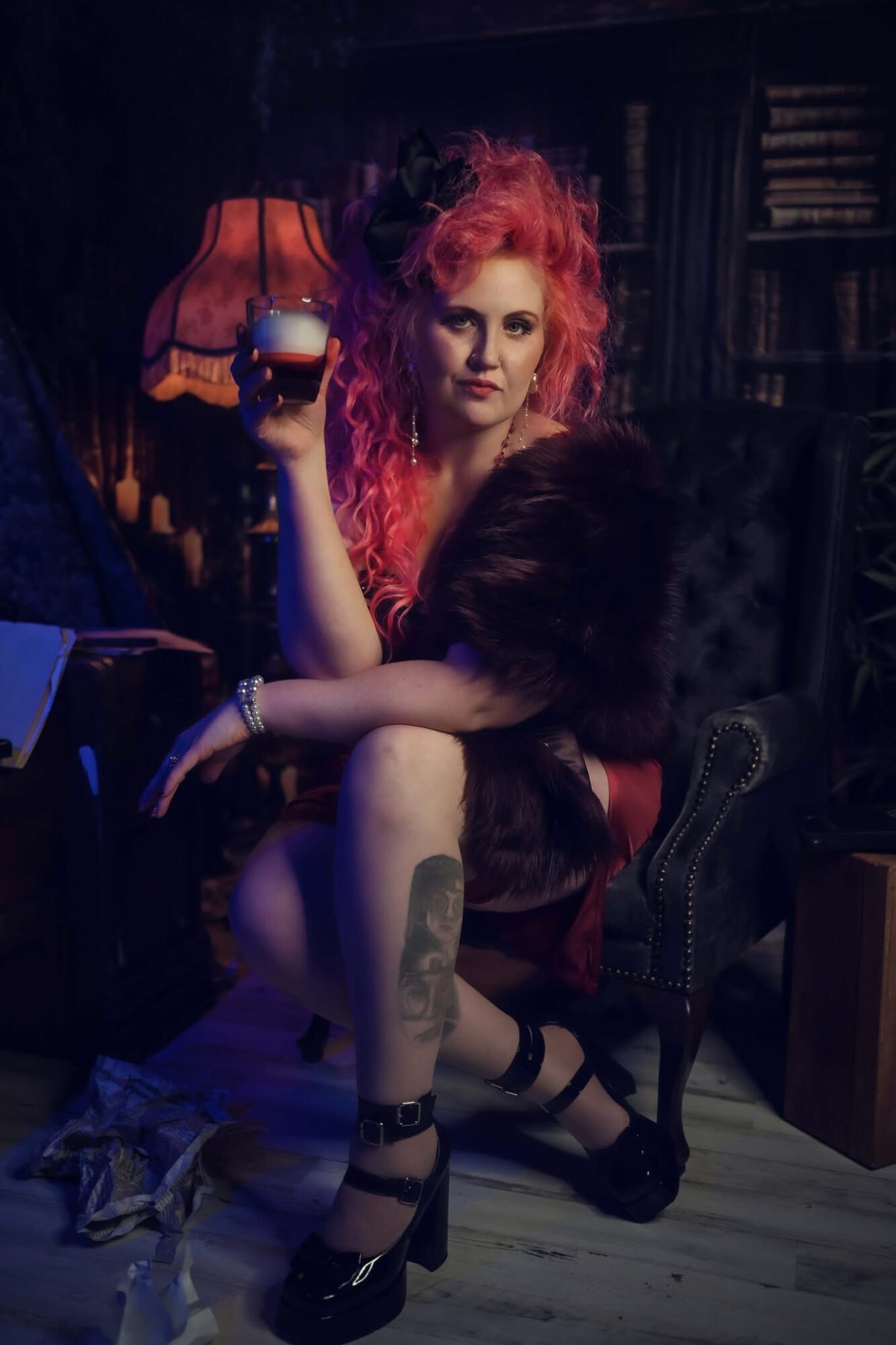 Woman with curly pink hair sitting in a dark room holding a glass, surrounded by books and vintage decor.