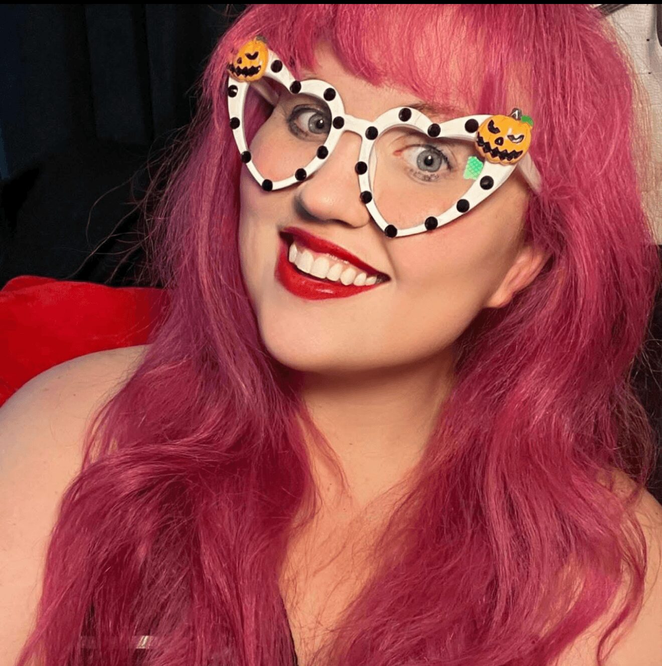 Woman with pink hair wearing Halloween-themed glasses with pumpkin decorations, smiling with red lipstick.