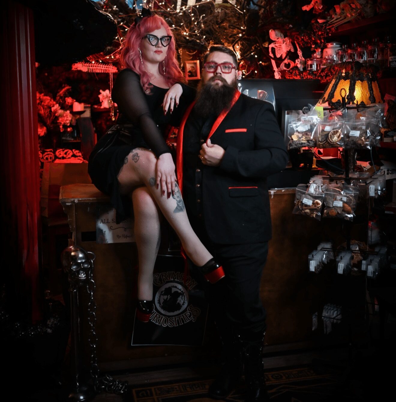 A woman with pink hair and glasses sitting on a bar counter with a man with a beard, both dressed stylishly, in a dimly lit bar.