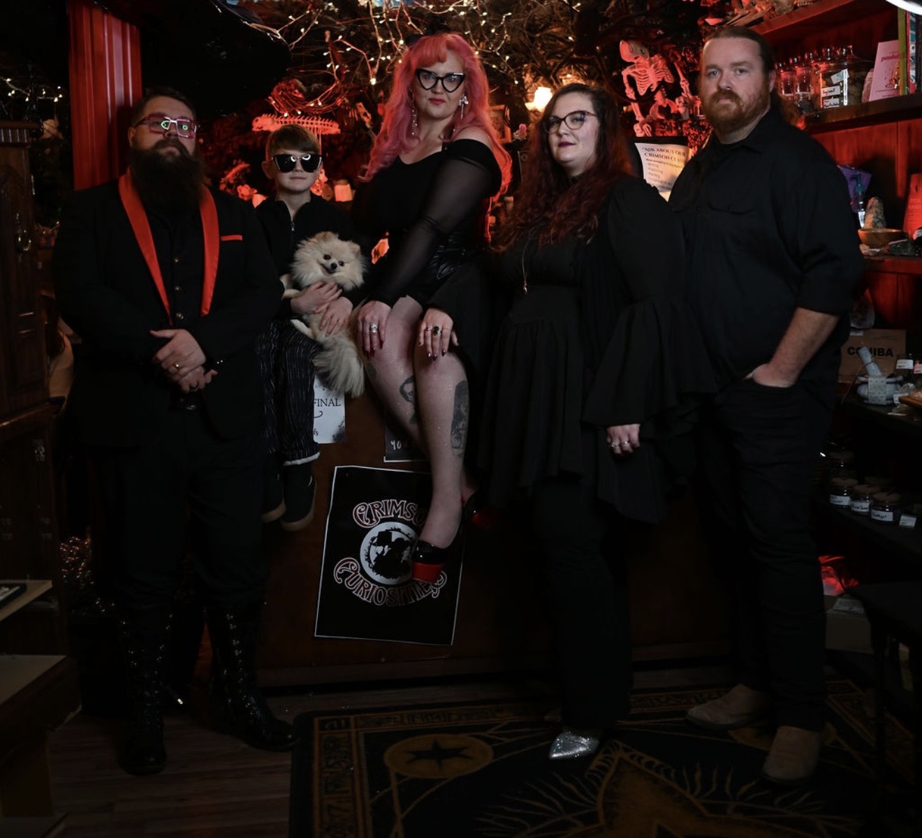 Group of five people and a dog in a dimly lit room with red lighting, some wearing dark clothing and glasses.