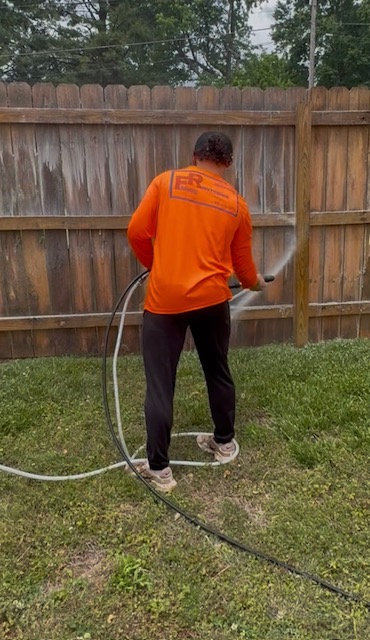 Person in orange jacket watering a wooden fence with a garden hose, standing on grass.