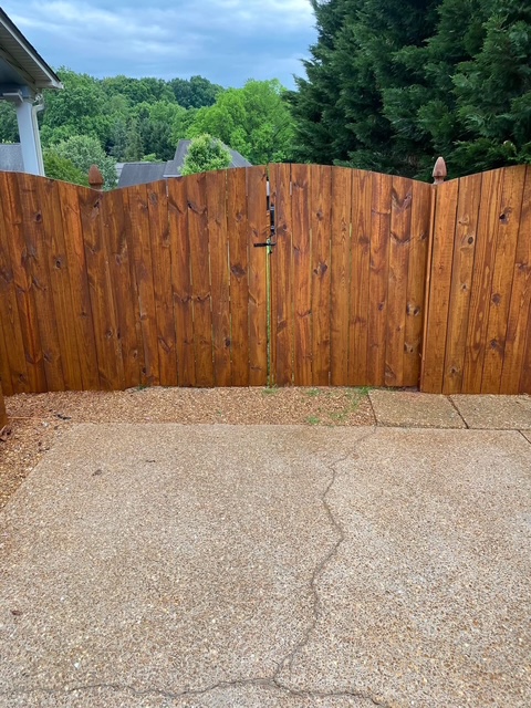 Wooden gate with a latch, part of a fence, with a concrete ground and green trees in the background.