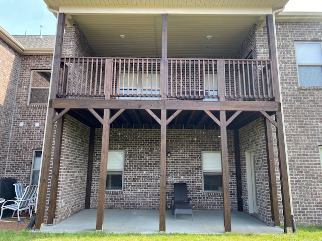 Two-story brick building with a wooden balcony and porch, supported by wooden posts, with chairs on the porch.