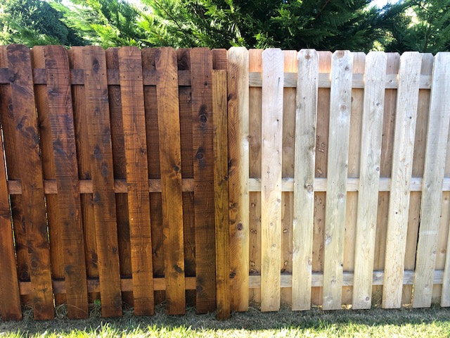Split wooden fence with dark brown on the left and light beige on the right, with green foliage above.